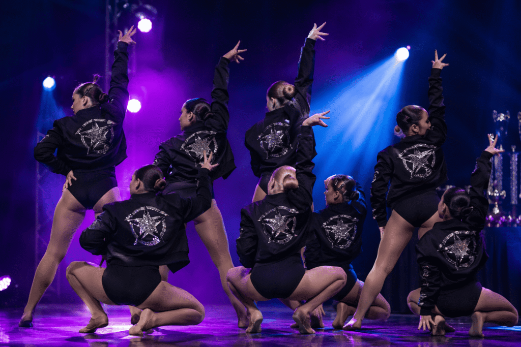 Dance team on competition stage wearing black cropped jackets with rhinestone back logos and sleeve names under dramatic purple spotlights — custom dance team jackets for competition performances and stage branding
