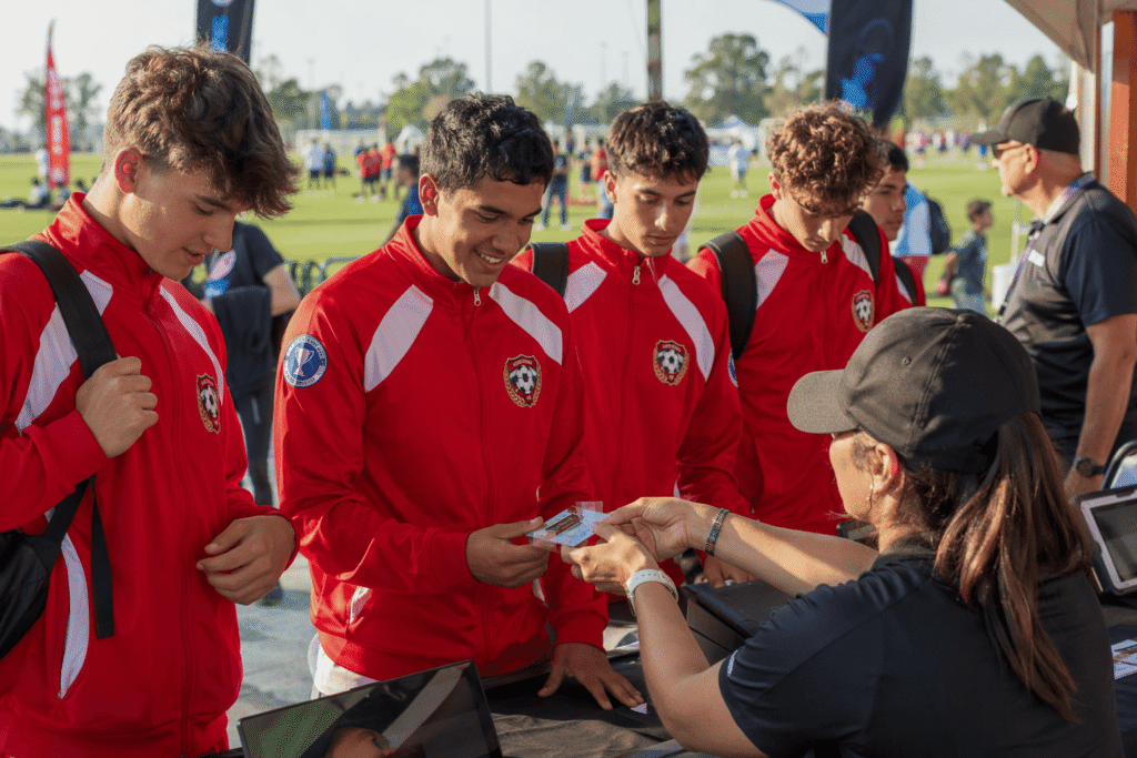 Sports tournament participants in bright red team jackets with embroidered tournament crest checking in at outdoor venue registration tent — custom event jackets for sports tournaments and competitive events