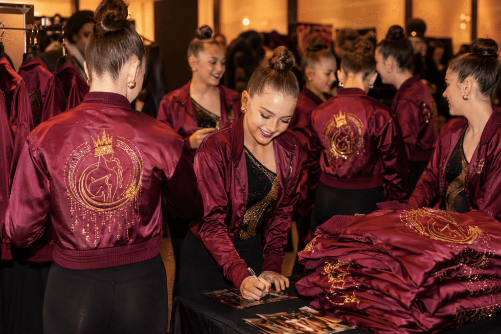 Dance team in burgundy satin jackets with gold embroidered logos at merchandise showcase event after performance — custom dance team jackets for team merchandise and fan apparel branding
