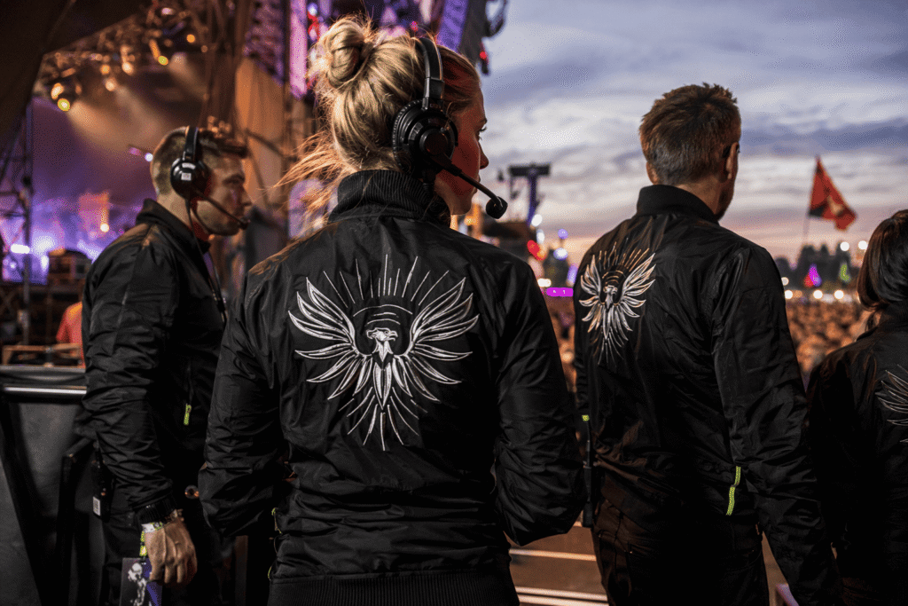 Music festival crew in black bomber jackets with metallic embroidered festival logo and bold back print working backstage at outdoor concert — custom event jackets for music festivals and production staff