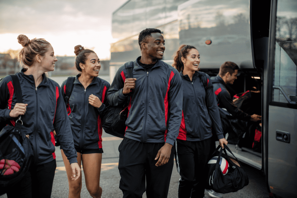 Multi-sport athletes in matching charcoal gray team jackets boarding charter bus, coordinated team travel scene — custom team jackets for multi-sport organizations and team trips