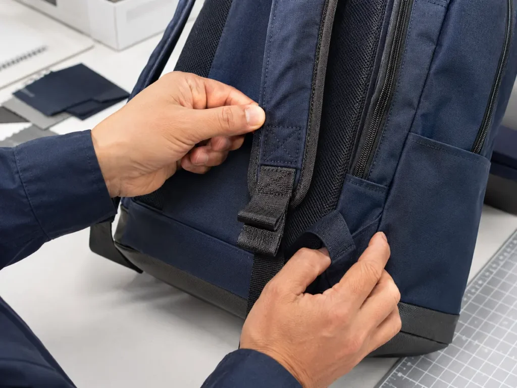 Technician checking reinforced stitching and shoulder-strap attachment on a navy backpack